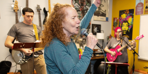 Members of PMAC's Alt Rock Band -- a female adult singer in the center holds a microphone in one hands with the other hand in the air; on the left is a male guitar player; on the right is a female bass player playing a pink bass.
