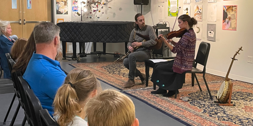 A fiddle player and bagpipe player sit playing in front of a row of audience members (adults and children)