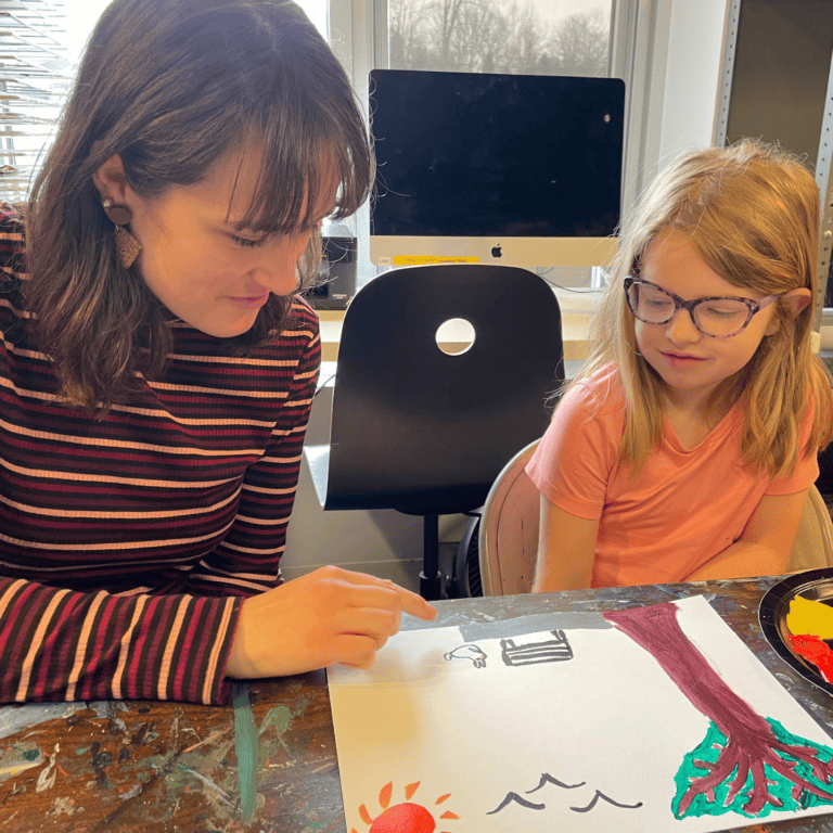 A private lesson teacher sits next to a young female student. They are both looking at a piece of artwork painted by the student that shows a tree, sun, birds in the sky, and a bunny on the ground.