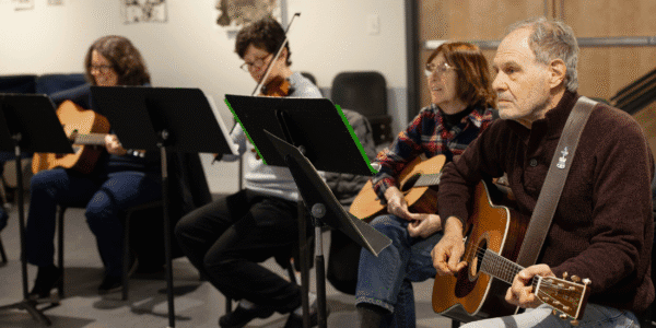 Guitars, fiddle, ukulele players play acoustically at music stands playing.