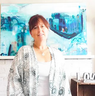 Artist Pamela Bates stands in front of a horizontal piece of artwork in teals and blues. She has brown hair pulled back and is smiling.