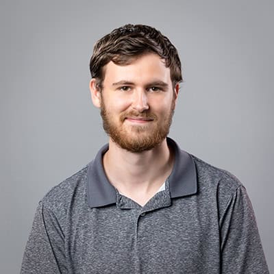 Eli Vannata in a dark gray collared shirt in front of a gray backdrop.