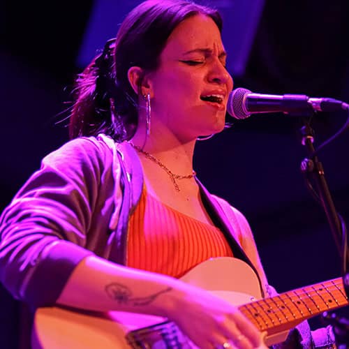 Kate Possi sings into a microphone while holding a tan guitar, lit by purple stage lights.