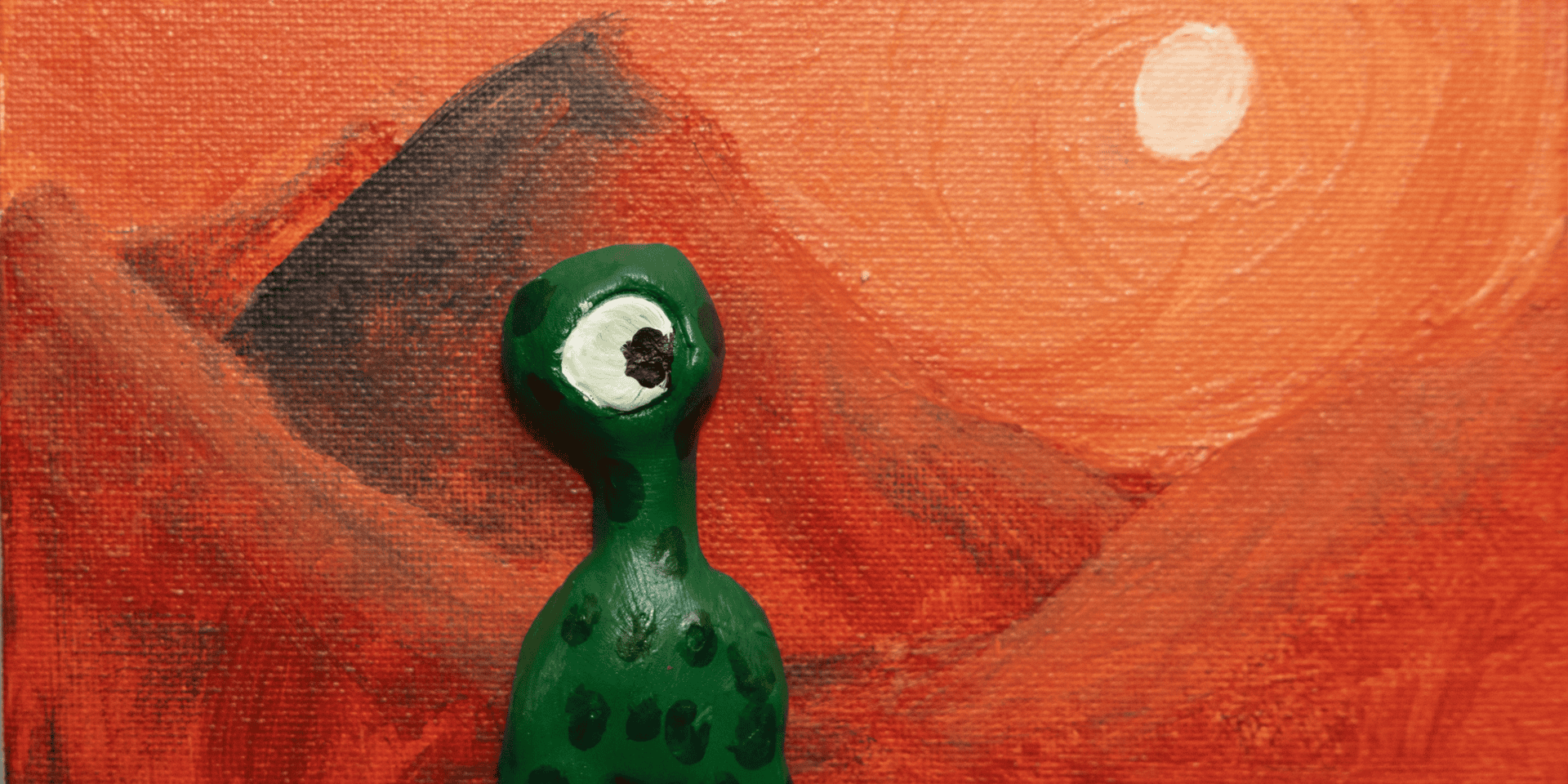 A piece of 3d artwork with a small green alien with 1 eye in front of a painted orange backdrop of dunes and a sun.