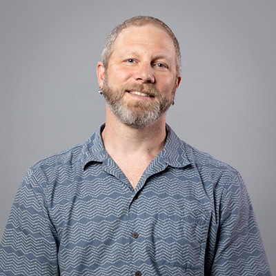 Rob Sanzone smiles in a blue collared shirt against a gray backdrop.