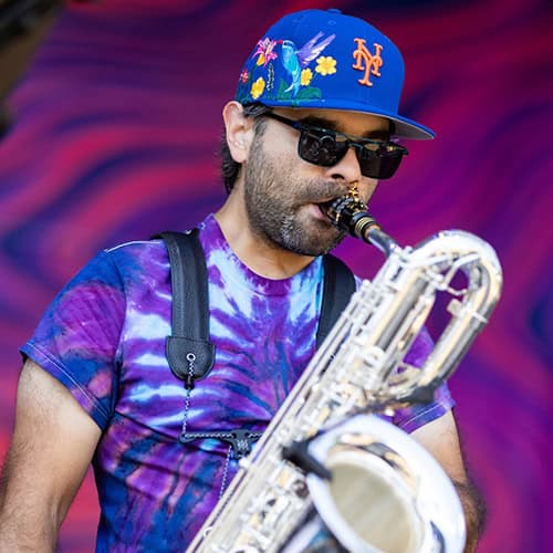 Will Cicola plays a silver bari saxophone while wearing a tie-dye shirt, blue hat, and sunglasses.