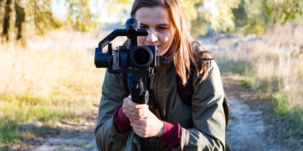 A teenage girl holds a gimbal with a mirrorless digital camera, against an outdoor background